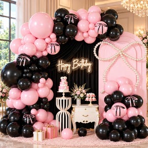 May include: A birthday party display featuring a large balloon arch in black and light pink, with a neon sign reading Happy Birthday. A white tiered cake and cupcakes sit on white pedestals. A pink arched backdrop is decorated with a pearl bow.