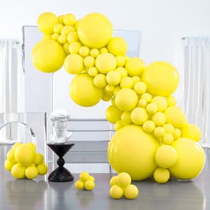 May include: A vibrant display of yellow balloons in various sizes, arranged in an arch and scattered on a surface. A white bust sculpture and a black and white pedestal add an artistic touch to the scene. The balloons create a festive and celebratory atmosphere.