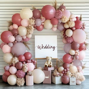 May include: A large arch of balloons in shades of dusty rose, cream, and mauve, accented with dried floral clusters. A white square sign with the word Wedding in dark script sits in the center, with a small plush teddy bear on a box below.
