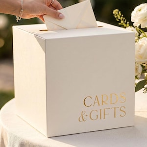 May include: A square, off-white card box featuring a horizontal slot on the top surface. The front panel displays the text "CARDS & GIFTS" in a metallic gold, serif font. The box is constructed with clean, sharp edges and a smooth, matte finish.