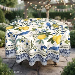 May include: A round tablecloth featuring a botanical print of yellow lemons, blue berries, green leaves, and blue birds on a white background. The bottom edge has a decorative border with a blue and white intricate pattern and the text pop joy party.