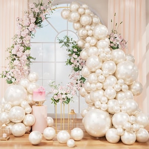 May include: A decorative party scene featuring a large balloon arch and clusters of pearl-like balloons in various sizes. Pink flowers and a small cake add to the festive atmosphere. The scene is set against a window with sheer curtains.