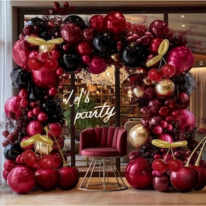 May include: A festive party scene with a burgundy velvet chair and a balloon arch. The arch is adorned with black, burgundy, and gold balloons, along with cherry and leaf accents. The neon sign says "Let's party".