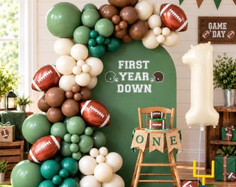 May include: A balloon arch in shades of olive green, brown, and cream, featuring football-shaped foil balloons and a green backdrop with the text FIRST YEAR DOWN. A wooden high chair displays a banner reading ONE with a small cake.
