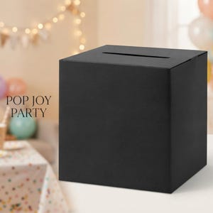 May include: A matte black square card box featuring a horizontal slot on the top surface for collecting cards or notes. The box has a clean, solid-colored exterior and is designed as a functional container for events or gatherings.
