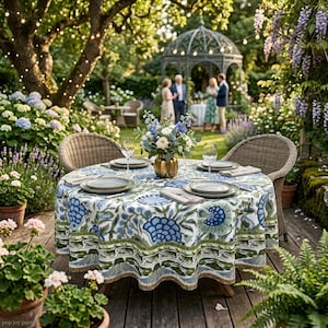 May include: A round dining table set on a wooden deck features a white tablecloth with a blue and green floral pattern. The table is set with four plates, four napkins, and a central gold vase filled with white and blue flowers.