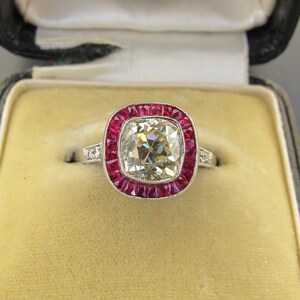 May include: A square-cut diamond ring set in platinum with a halo of red rubies. The ring is in a black velvet ring box with a white satin lining. The box has a label that reads "C. Barrald, Antique."