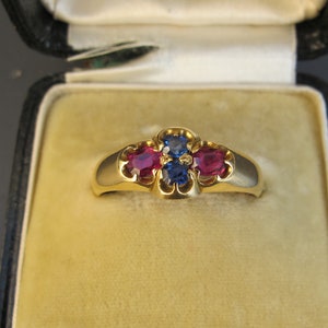 May include: A gold ring with a central blue gemstone flanked by two red gemstones, set in a decorative setting. The ring is displayed in a black jewelry box with a gold clasp and a yellow velvet interior.