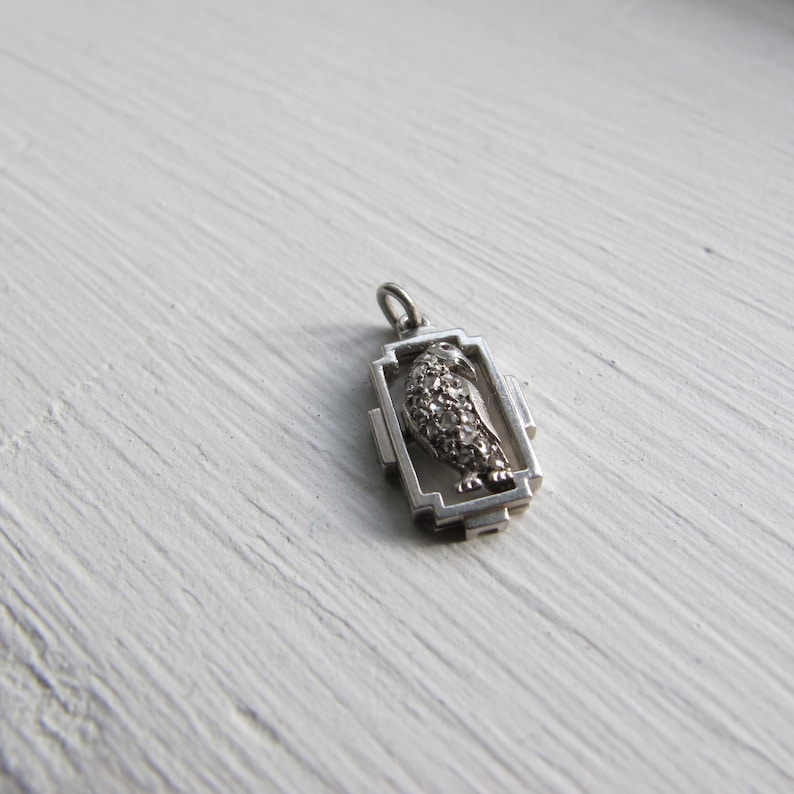 May include: A silver-toned pendant featuring a bird design. The bird is detailed with small, sparkling accents, possibly gemstones, within a rectangular frame. The pendant has a small loop for attaching to a chain or bracelet.