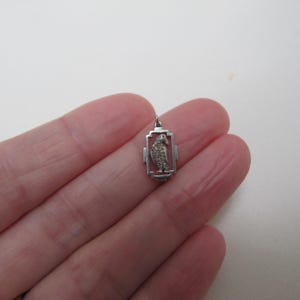May include: A small, silver-toned pendant featuring a bird design within a rectangular frame. The bird is detailed with textured elements, and the pendant has a small loop for a chain. The pendant is held in a person's hand.