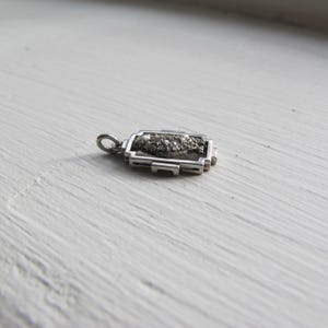 May include: A small, rectangular silver pendant with a cluster of small, dark stones in the center. The pendant has a geometric design with a loop at the top for a chain. The pendant is lying on a white surface.