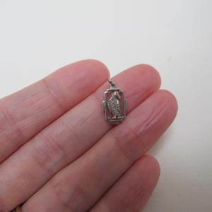 May include: A small, silver-toned pendant featuring a detailed bird design within a rectangular frame. The bird has textured detailing, and the frame has a cross-like design on each side. The pendant is attached to a small loop for a chain.