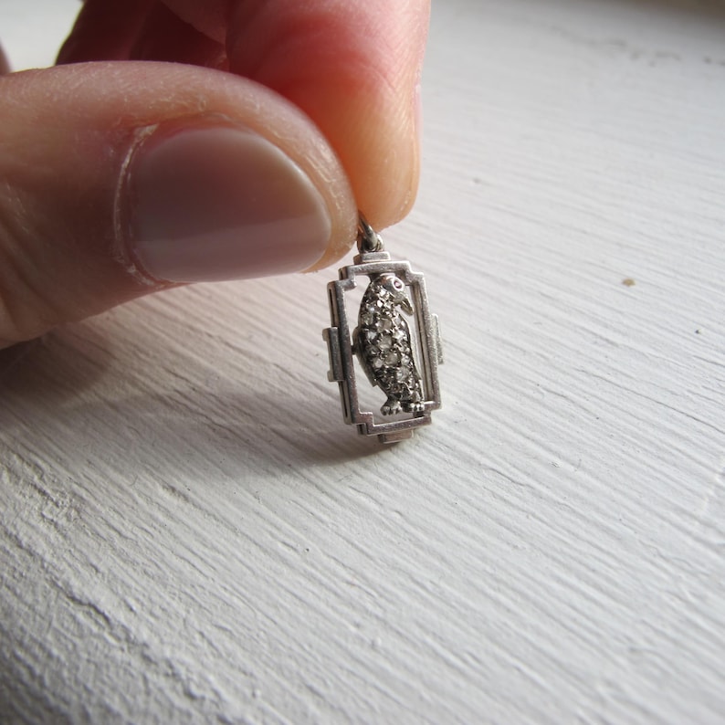 May include: A small, silver-toned pendant featuring a bird design encrusted with small, sparkling stones. The bird is set within a rectangular frame with a geometric design. The pendant is held up by a small loop for a chain.