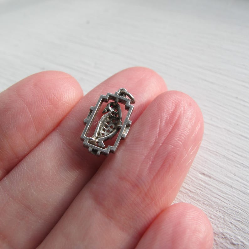 May include: A small, silver-toned pendant featuring a stylized fish design within a rectangular frame. The fish has a textured, scale-like pattern. The pendant is likely made of metal and is designed to be worn as jewelry.
