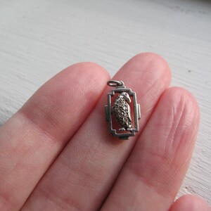 May include: A small, silver-toned pendant featuring a bird design within a rectangular frame. The bird's body is detailed with small, textured elements, and the pendant has a loop for attaching to a chain. The pendant is held in a hand.
