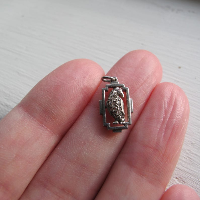 May include: A small, silver-toned pendant featuring a bird design within a rectangular frame. The bird's body is detailed with small, textured elements, and the pendant has a loop for attaching to a chain. The pendant is held in a hand.