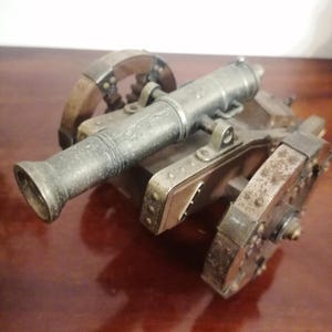 May include: An antique metal cannon model with a dark, aged finish. The miniature artillery piece features a barrel, carriage, and spoked wheels, showcasing intricate detailing and rivets. The cannon is a historical replica, reminiscent of a bygone era.