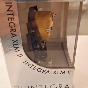 Può includere: Una testina fonografica ADC Integra XLM II con corpo color oro alloggiata in una scatola espositiva in plastica trasparente. La confezione riporta il testo INTEGRA XLM II e Audio Dynamics Corporation stampato in nero sulla base e sui lati.
