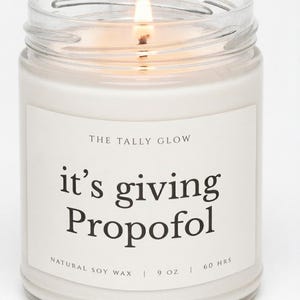 Op de afbeelding: Een kaars in een doorzichtige glazen pot met een brandende lont en witte was. Het etiket vermeldt THE TALLY GLOW, it's giving Propofol, NATURAL SOY WAX, 9 OZ, 60 HRS. De kaars geeft wat rook af en het glas heeft een zichtbare rand.
