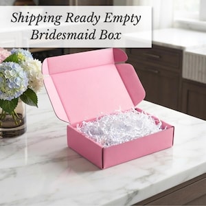 May include: A light pink cardboard box with an open lid, filled with white shredded paper filler. The box is rectangular and features a hinged top flap. The text Shipping Ready Empty Bridesmaid Box is displayed above the box in a black font.