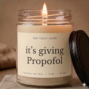 May include: A lit candle in a glass jar with a black lid. The label reads "THE TALLY GLOW" and "it's giving Propofol." The candle is made of natural soy wax and is 255 grams.