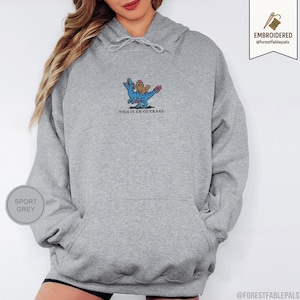 May include: Sport grey hoodie with a kangaroo pocket and drawstring hood. The front features an embroidered design of a blue dinosaur carrying a brown creature, with the text "THIS IS AN OUTRAGE" below. The brand name is embroidered on the upper right.