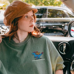 Może przedstawiać: Koszulka w kolorze mchu z kreskówkowym dinozaurem i napisem "THIS IS AN OUTRAGE". Zestaw uzupełniają podarte czarne dżinsy, brązowa sztruksowa czapka i białe trampki.