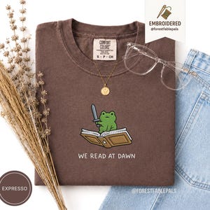 以下が含まれることがあります： ダークブラウンのTシャツで、灰色の剣を持ち、開いたベージュの本の上に座っている緑色のカエルの刺繍が施されています。刺繍の下には「WE READ AT DAWN」という文字があります。Tシャツの首元にはComfort Colorsのラベルが付いています。