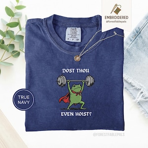 May include: Navy blue t-shirt featuring a cartoon frog lifting weights. The frog is wearing a red cape and a green bow tie. The text on the shirt reads "DOST THOU EVEN HOIST?" and "TRUE NAVY". The t-shirt is from Comfort Colors.