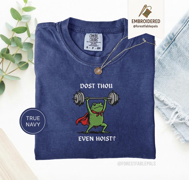 May include: Navy blue t-shirt featuring a cartoon frog lifting weights. The frog is wearing a red cape and a green bow tie. The text on the shirt reads "DOST THOU EVEN HOIST?" and "TRUE NAVY". The t-shirt is from Comfort Colors.