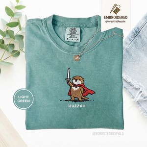 May include: Light green t-shirt with an embroidered otter design. The otter is wearing a red cape and holding a sword, with the word "Huzzah" below. A silver necklace with a pendant is also visible.