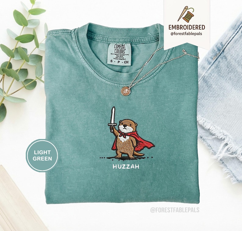 May include: Light green t-shirt with an embroidered otter design. The otter is wearing a red cape and holding a sword, with the word "Huzzah" below. A silver necklace with a pendant is also visible.