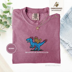 May include: Berry-colored t-shirt with an embroidered design of a blue dinosaur carrying a cat wearing sunglasses and a crown. The text "MONGO IS APPALLED" is below the design. A gold necklace is draped across the shirt.