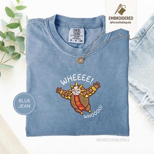 Könnte beinhalten: Ein blaues Comfort Colors T-Shirt mit einem gestickten Cartoon-Charakter-Design. Das Design zeigt eine braune und goldene Figur mit den Worten "WHEEEE! WHOOOO!" in Weiß. Eine goldene Halskette ist über dem Shirt drapiert.