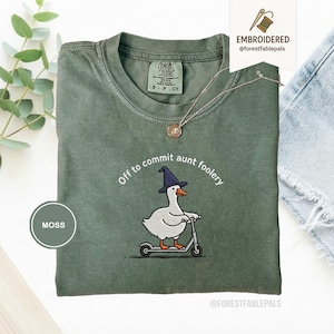Puede incluir: Camiseta Comfort Colors verde musgo con la frase bordada "Off to commit aunt foolery". El diseño presenta un pato de dibujos animados con un sombrero de bruja montado en un scooter. Un collar plateado está sobre la camiseta.