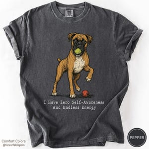 May include: A charcoal grey t-shirt featuring a graphic illustration of a brown and white boxer dog holding a green tennis ball in its mouth, with a red ball on the ground and the text "I Have Zero Self-Awareness And Endless Energy" printed below.