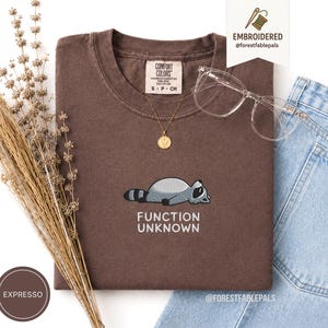 Op de afbeelding: Een bruin Comfort Colors t-shirt met een geborduurd wasbeerontwerp en de tekst "FUNCTION UNKNOWN". Het shirt wordt gecombineerd met een gouden ketting, een doorzichtige bril en een spijkerbroek. De kleur van het shirt is "Expresso".