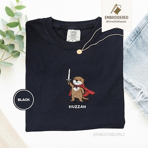May include: Black t-shirt with an embroidered otter design. The otter is wearing a red cape and holding a sword. The word "Huzzah" is embroidered below the otter. A gold necklace is draped on the shirt.