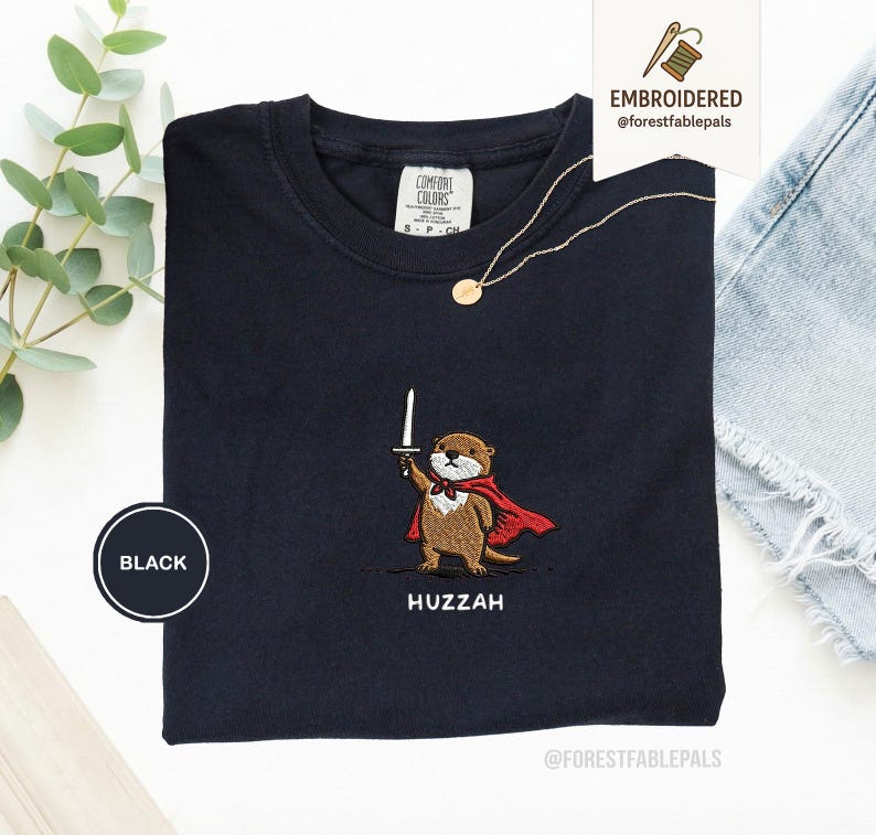May include: Black t-shirt with an embroidered otter design. The otter is wearing a red cape and holding a sword. The word "Huzzah" is embroidered below the otter. A gold necklace is draped on the shirt.
