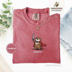 May include: Crimson-colored t-shirt with an embroidered otter design. The otter is depicted holding a sword and wearing a red cape, with the word "Huzzah" below. The shirt has a crew neck and short sleeves.