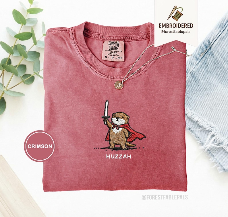 May include: Crimson-colored t-shirt with an embroidered otter design. The otter is depicted holding a sword and wearing a red cape, with the word "Huzzah" below. The shirt has a crew neck and short sleeves.
