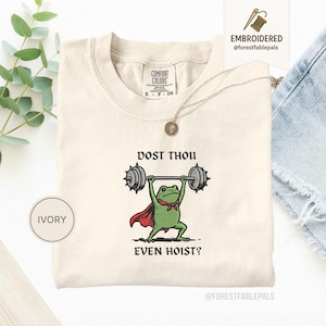 May include: Ivory t-shirt with a cartoon frog lifting weights, the text reads "DOST THOU EVEN HOIST?". The frog is wearing a red cape and a green bow tie. The shirt has a crew neck and short sleeves. A gold necklace is draped across the shirt.