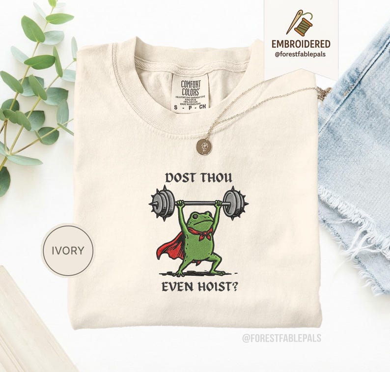 May include: Ivory t-shirt with a cartoon frog lifting weights, the text reads "DOST THOU EVEN HOIST?". The frog is wearing a red cape and a green bow tie. The shirt has a crew neck and short sleeves. A gold necklace is draped across the shirt.