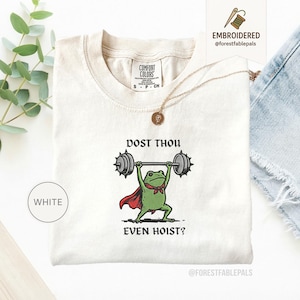 May include: Cream-coloured t-shirt with an embroidered design of a green frog lifting weights. The frog wears a red cape and a green bow tie. The text "DOST THOU EVEN HOIST?" is above and below the frog. The shirt has a crew neck and short sleeves.