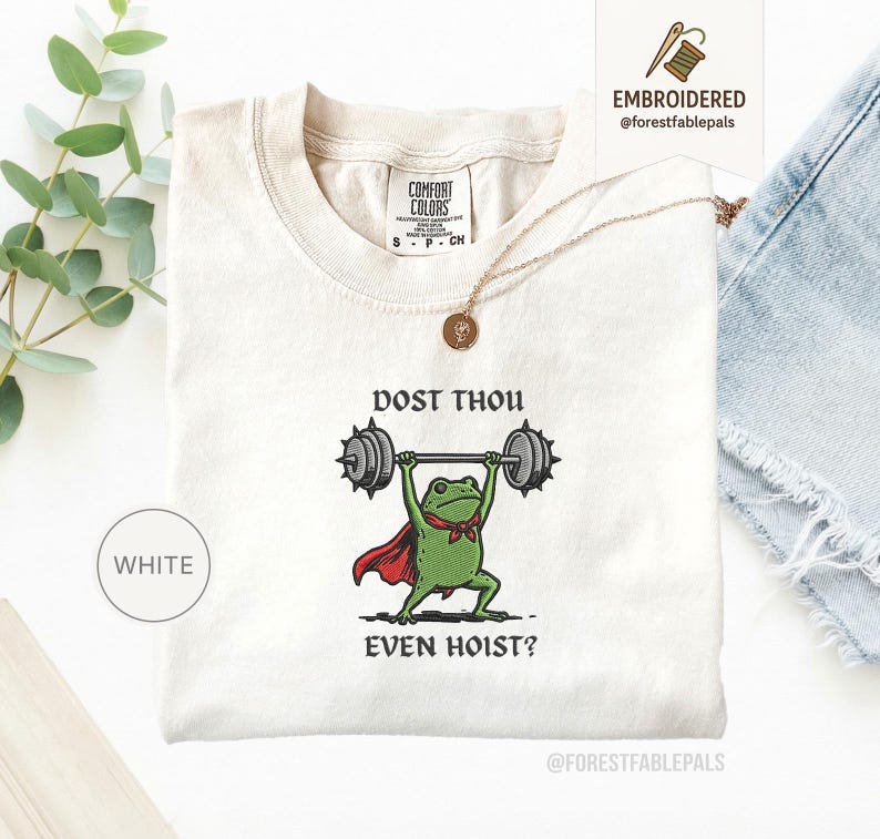 May include: Cream-coloured t-shirt with an embroidered design of a green frog lifting weights. The frog wears a red cape and a green bow tie. The text "DOST THOU EVEN HOIST?" is above and below the frog. The shirt has a crew neck and short sleeves.