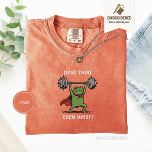 May include: Orange Comfort Colors t-shirt featuring a green frog lifting weights, wearing a red cape and a bow tie. The text "DOST THOU EVEN HOIST?" is above and below the frog. A silver necklace is on the shirt.