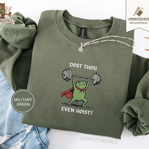 Puede incluir: Sudadera verde militar con un diseño bordado de una rana levantando pesas, con una capa roja y una pajarita. El texto "DOST THOU EVEN HOIST?" está encima de la rana. Un collar plateado está sobre la sudadera.