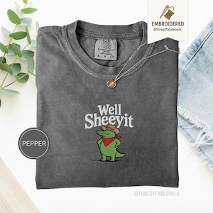 May include: A charcoal grey t-shirt featuring an embroidered graphic of a green alligator wearing a brown cowboy hat and a red bandana. The text "Well SHEEYIT" is embroidered in white above the alligator. The shirt has a crew neckline.