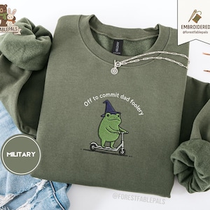 Op de afbeelding: Een olijfgroene sweatshirt met de geborduurde tekst "Off to commit dad foolery" en een cartoonkikker op een scooter. De kikker draagt een heksenhoed. Een zilveren ketting is over de sweatshirt gedrapeerd.