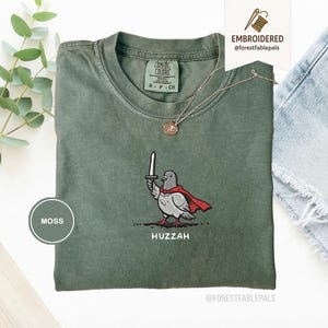 Op de afbeelding: Mosgroen T-shirt met een geborduurd ontwerp van een duif met een rode cape en een zwaard. Het woord "Huzzah" is eronder geborduurd. Het shirt heeft een ronde hals en korte mouwen.